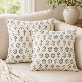 Two square throw pillows with a repeating navy blue damask pattern on a cream background, styled on a beige sofa with a knit blanket and a potted plant in the background.