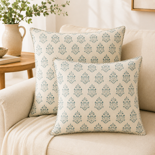 Two square throw pillows with a repeating blue-gray damask pattern on a cream background, styled on a beige sofa in a living room setting.