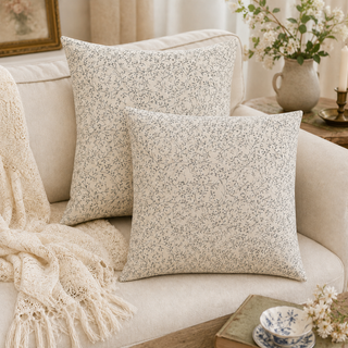 Two square throw pillows with a delicate beige and white floral print are styled on a cream-colored sofa, alongside a matching cream knit throw blanket and a cozy, warm home interior setting.