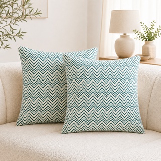 Two square throw pillows with a teal and cream chevron pattern resting on a beige sofa in a bright, modern living room setting.