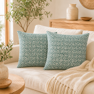 Two square throw pillows with a small-scale floral pattern in teal and cream, styled on a cream-colored sofa with a beige knit throw blanket in a warm, sunlit living room.